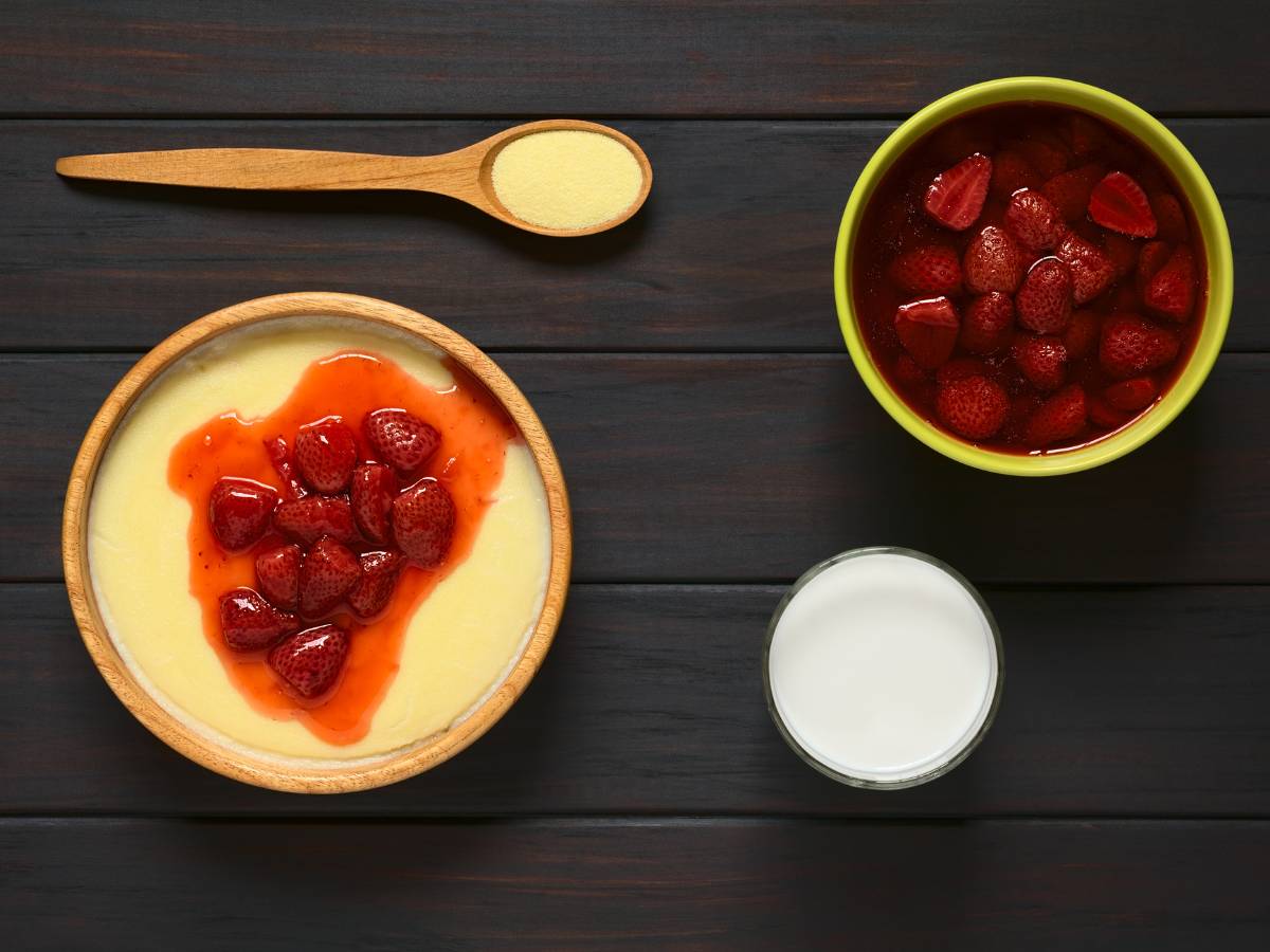 Na ciemnym tle, stoją miski z kaszą manną, polaną ugotowanymi truskawkami, miseczka z gotowanymi truskawkami, miseczka ze śmietaną i leży łyżka z suchą kaszą manną. Ilustracja do artykułu: "Grysik a kasza manna - porównanie wartości odżywczych i zastosowań w kuchni"