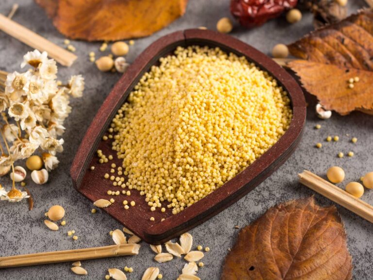Drewniane naczynie z żółtą kaszą, a obok rozsypane ziarna i listki suche. Ilustracja do artykułu: "Kasza jaglana a kuskus - porównanie wartości odżywczych i zastosowania w kuchni"