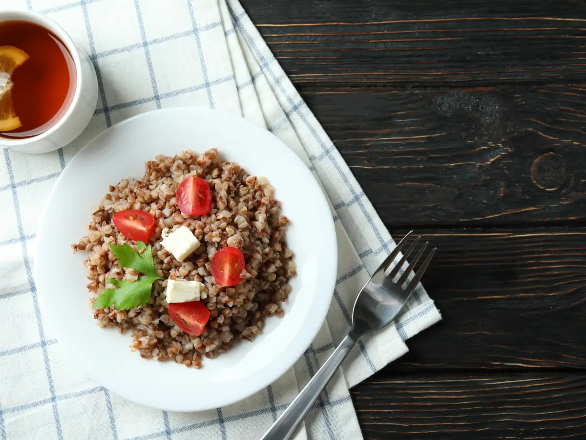 Talerz z kaszą i warzywami z serem na wierzchu, ustawiony na białym obrusie na ciemnym stole. Ilustracja do artykułu Dlaczego kasza gryczana na kolację w trakcie odchudzania to strzał w dziesiątkę?