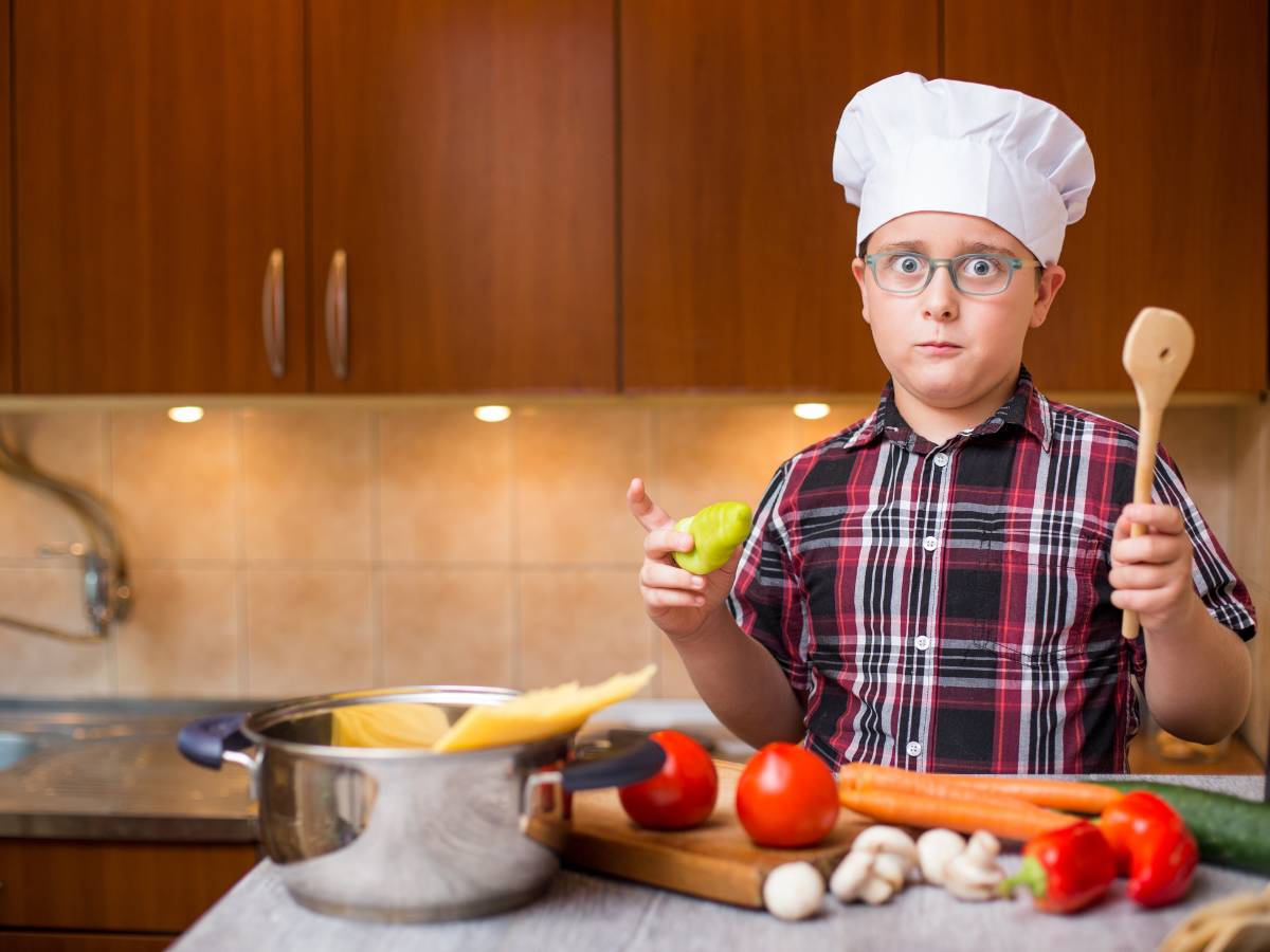 Chłopczyk w kucharskiej czapce, z drewnianą łyżką w dłoni przed stołem z warzywami i garnkiem. Ilustracja do artykułu: Odkryj 6 kreatywnych sposobów na to, co zrobić z ugotowanej kaszy jęczmiennej, abyś nie musiał wyrzucać