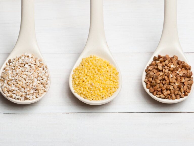 Trzy duże białe plastikowe łyżki na błękitnym tle z 3 rodzajami kaszy. Ilustracja do artykułu: "Sprawdź, czy kasza tuczy i jaki może mieć wpływ na wagę"