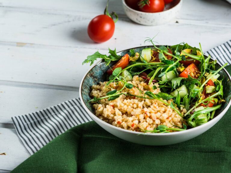 Na ciemnozielonym obrusie i drugim - w biało- granatowe paski - stoi miska bladoniebiedska z kaszą i sałatą z pomidorkami. Ilustracja do artykułu: "6 pomysłów na to, czym zastąpić kaszę bulgur w codziennym gotowaniu"