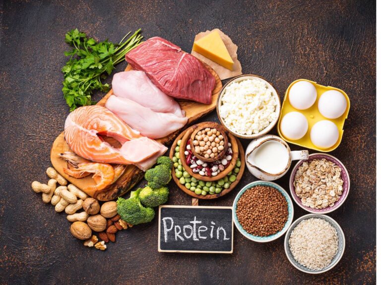 Napis Protein i w koło ułożone produkty białkowe: mięso, jajka, kasze w miseczkach, warzywa. Ilustracja do artykułu: Odkryj, która kasza ma najwięcej białka i buduj mięśnie