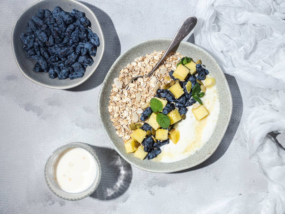 Na błękitnym obrusie , stoją - widziane od góry- kubek z jogurtem, miseczka jagód i talerz z suchymi płatkami, zieleniną i czyms białym. Ilustracja do artykułu: "5 pomysłów na to, z czym jeść płatki owsiane, aby były jeszcze smaczniejsze"