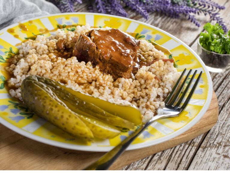 Biały talerz z kaszą i gulaszem oraz ogórkami. Ilustracja do artykuły "Odkryj 7 pysznych pomysłów na to, z czym podawać kaszę jęczmienną na obiad"